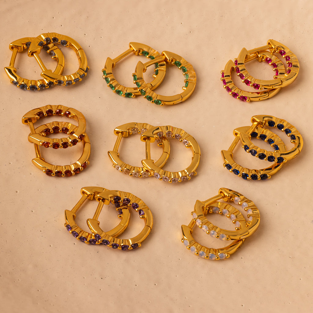 Nine pairs of gold hoop earrings, including Inside Out Blue Zircon Huggies with luminous gemstones, are arranged on a beige surface.