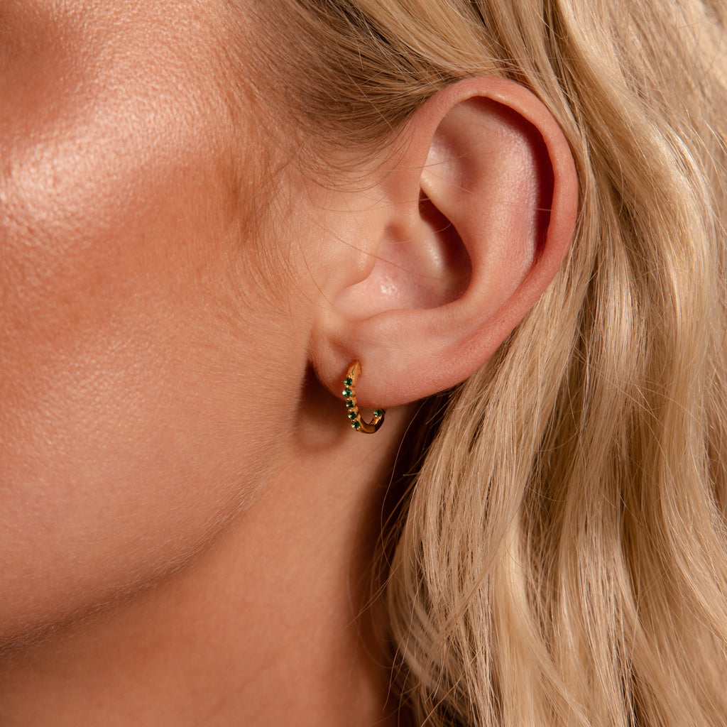 Close-up of a woman's ear with blonde hair wearing Inside Out Peridot Huggies—small gold hoop earrings with green stones, perfect for stacking or as elegant birthstone huggie earrings.