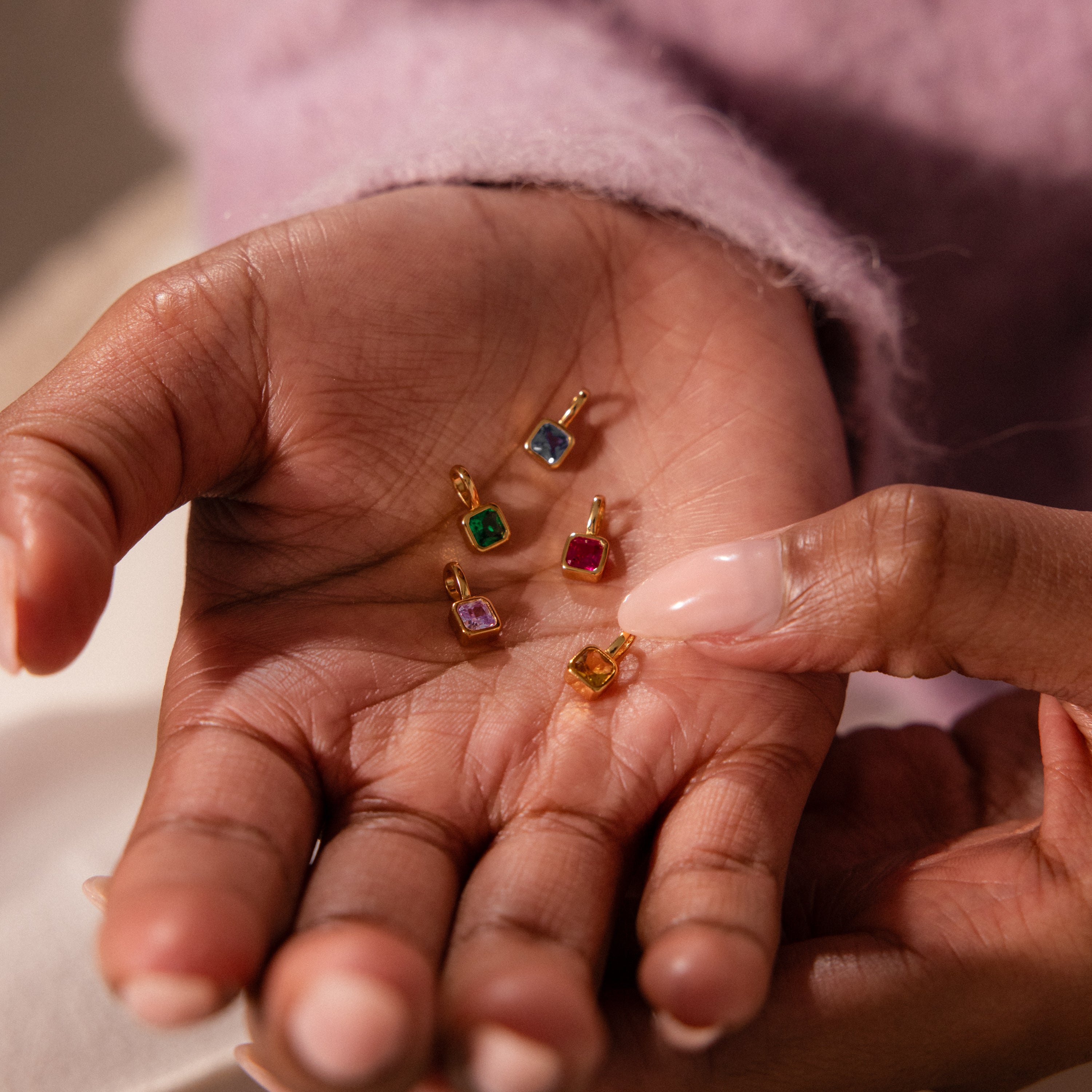 A hand holds four small, colorful gemstone pendants with gold frames, while another hand points to the Square Bezel Emerald Charm.
