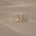 The Square Bezel Diamond Charm, a gold pendant featuring a square diamond, rests on a beige, softly textured background—an elegant piece for any gemstone jewelry collection.