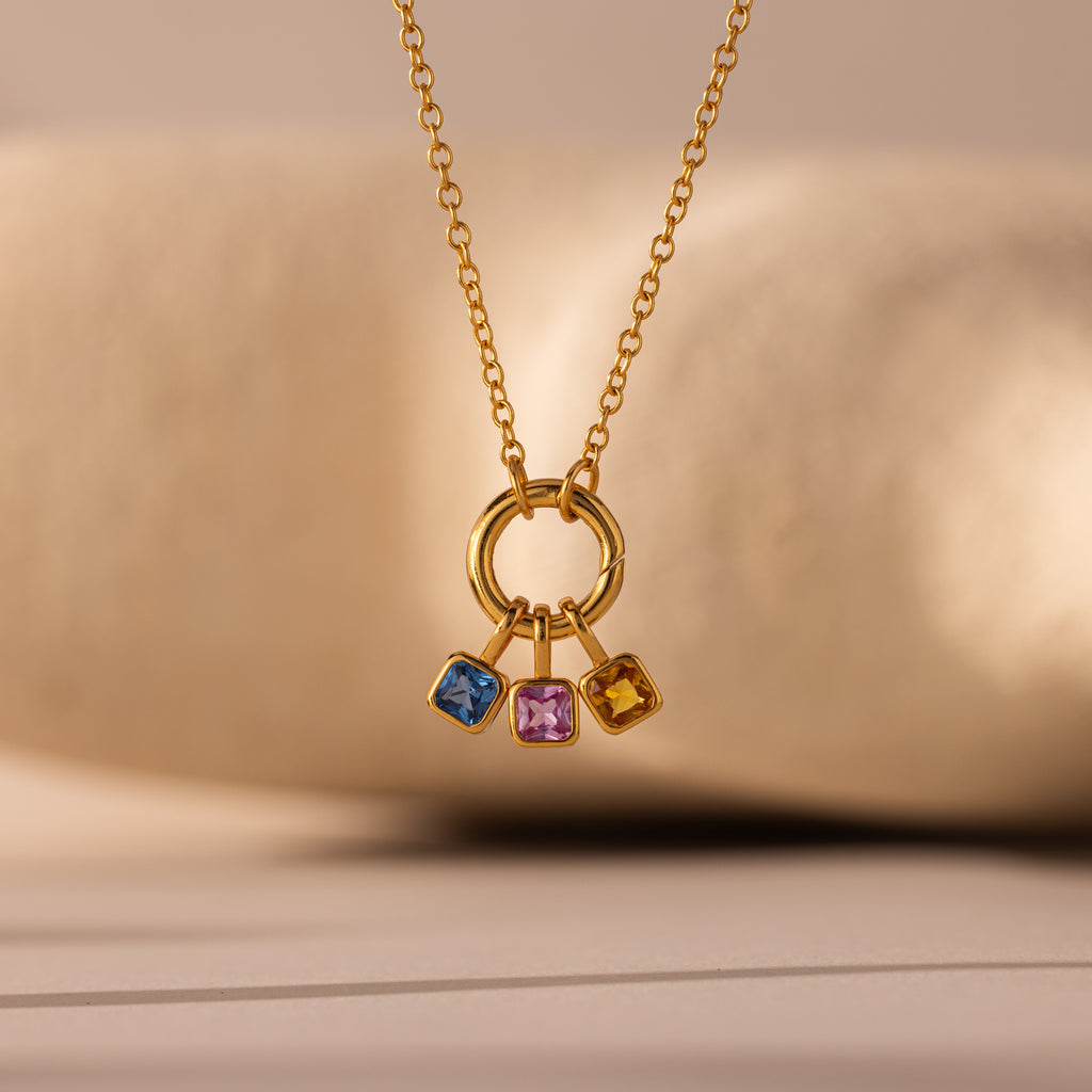 The Square Bezel Diamond Charm features a gold necklace with a circular pendant holding three square gemstones—blue, pink, and yellow—for a vibrant gemstone jewelry look.