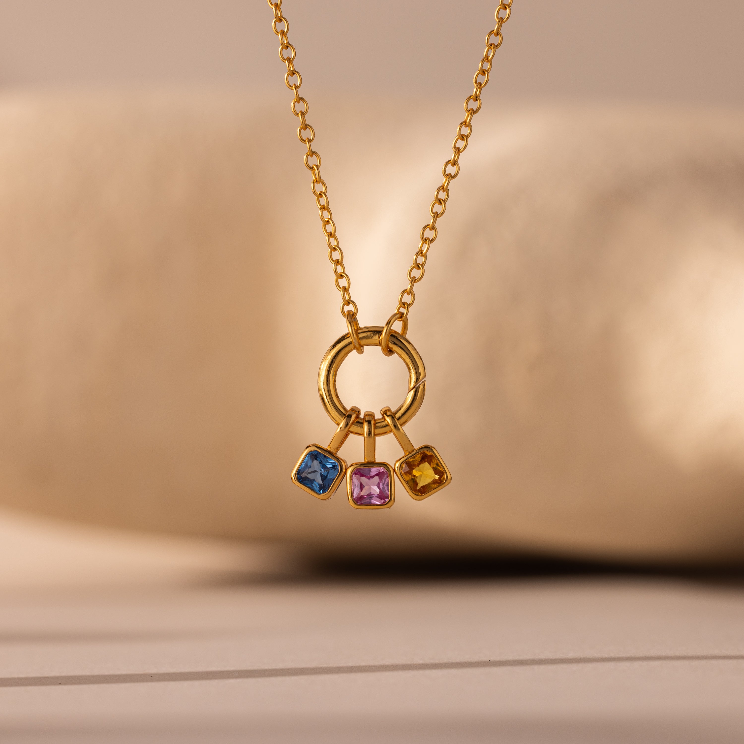 A gold necklace with a circular pendant showcases three Square Bezel Amethyst Charms—blue, pink, and yellow—making it an ideal choice for gemstone jewelry lovers.