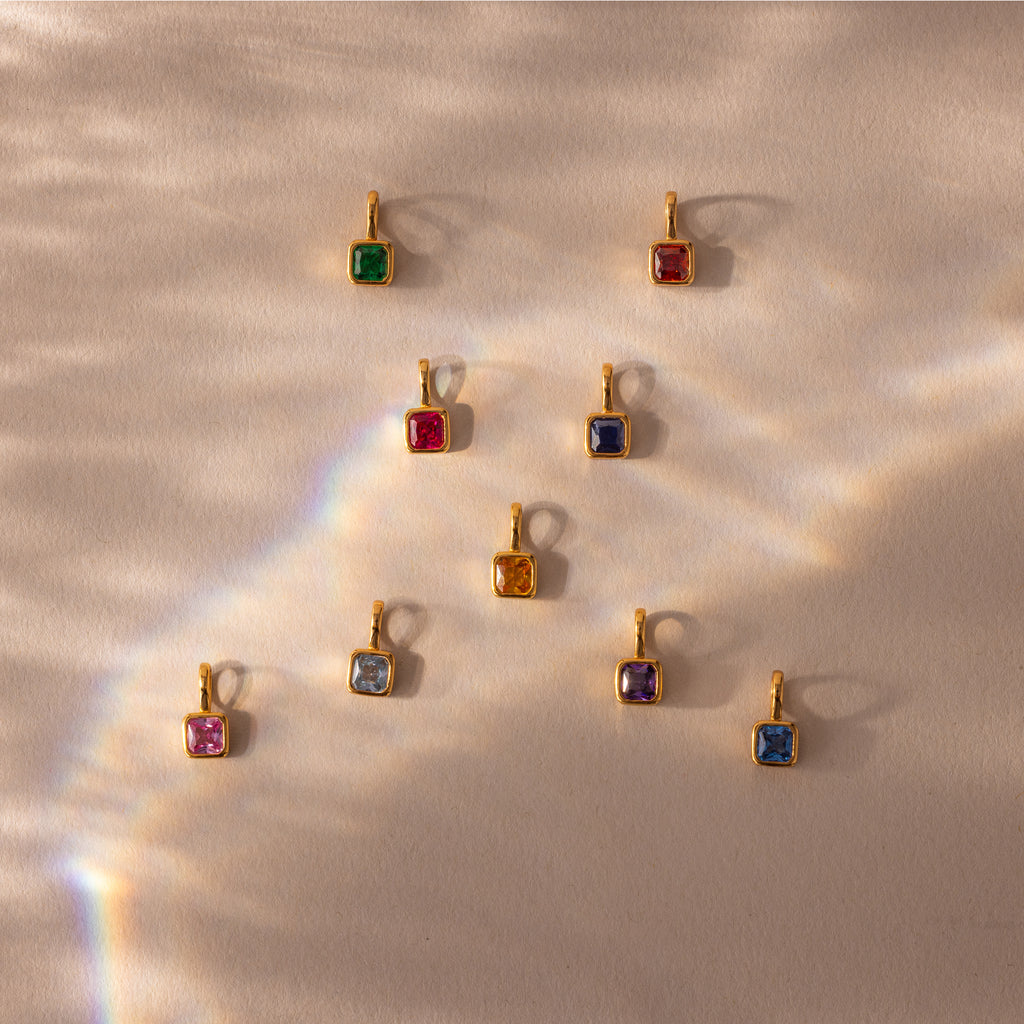 Nine Square Bezel Amethyst Charms with gold frames are arranged on a beige background, illuminated by soft light and rainbow reflections.