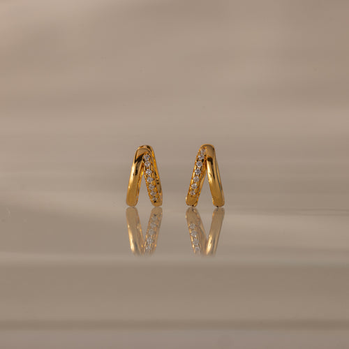 Amelia Duo Diamond Huggies – gold V-shaped modern earrings featuring sparkling diamonds, showcased on a reflective beige surface.
