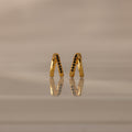 The Amelia Duo Onyx Huggies are modern V-shaped earrings featuring black onyx stones, elegantly reflected on a smooth, beige surface.