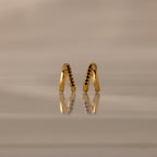 The Amelia Duo Onyx Huggies are modern V-shaped earrings featuring black onyx stones, elegantly reflected on a smooth, beige surface.