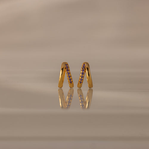 The Amelia Duo Amethyst Huggies, gold intertwined hoops with purple stones, are displayed upright on a reflective beige surface, adding a vibrant touch to your collection.
