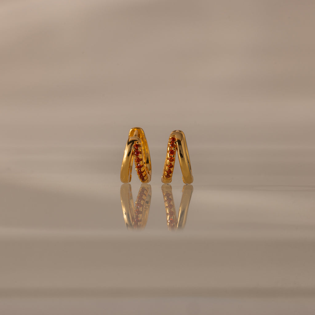 The Amelia Duo Garnet Huggies, featuring intertwined hoops and shimmering red gemstone accents, stand upright on a reflective beige surface.
