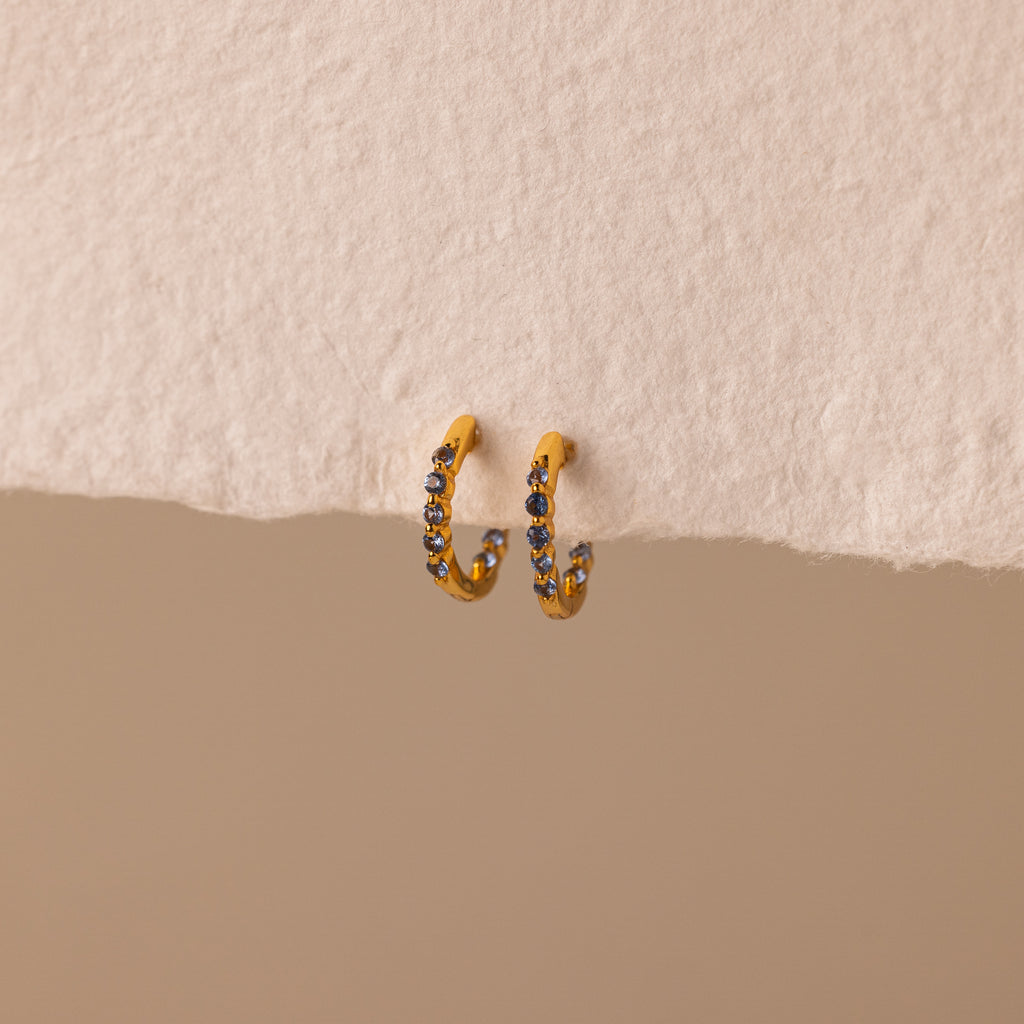 Inside Out Blue Zircon Huggies—small gold hoops adorned with luminous blue zircon gemstones, perfect as Birthstone Huggie Earrings—are displayed against a textured beige background.