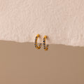 Inside Out Blue Zircon Huggies—small gold hoops adorned with luminous blue zircon gemstones, perfect as Birthstone Huggie Earrings—are displayed against a textured beige background.