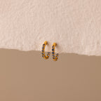 Inside Out Blue Zircon Huggies—small gold hoops adorned with luminous blue zircon gemstones, perfect as Birthstone Huggie Earrings—are displayed against a textured beige background.