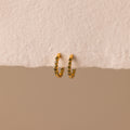 Inside Out Peridot Huggies are gold hoop stacking earrings featuring small green gemstones, perfect as birthstone huggie earrings. Shown on a textured off-white surface with a beige background.