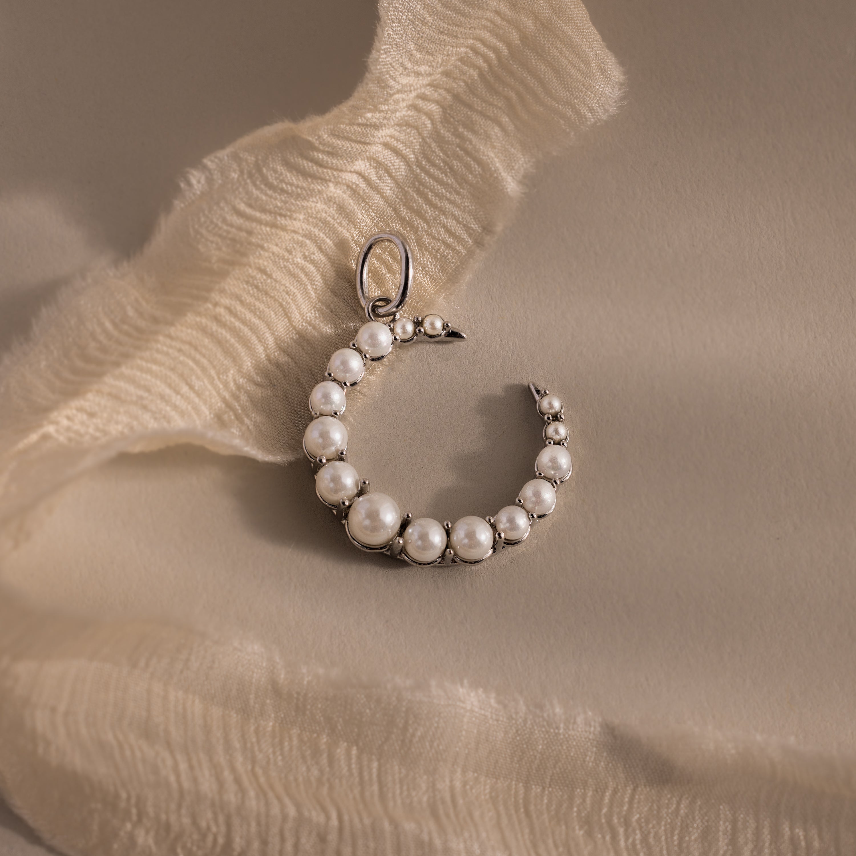 The Pearl Moon Charm, a crescent-shaped pearl earring inspired by celestial beauty, is displayed on soft cream-colored fabric.