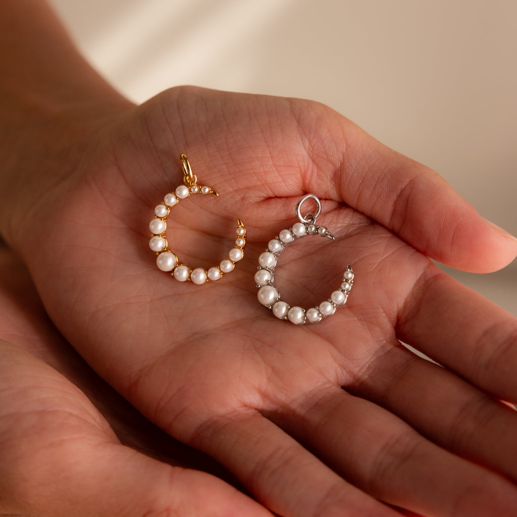 A hand holds two Pearl Moon Charm earrings—one gold, one silver—showcasing celestial beauty with their elegant crescent silhouette and luminous pearls.