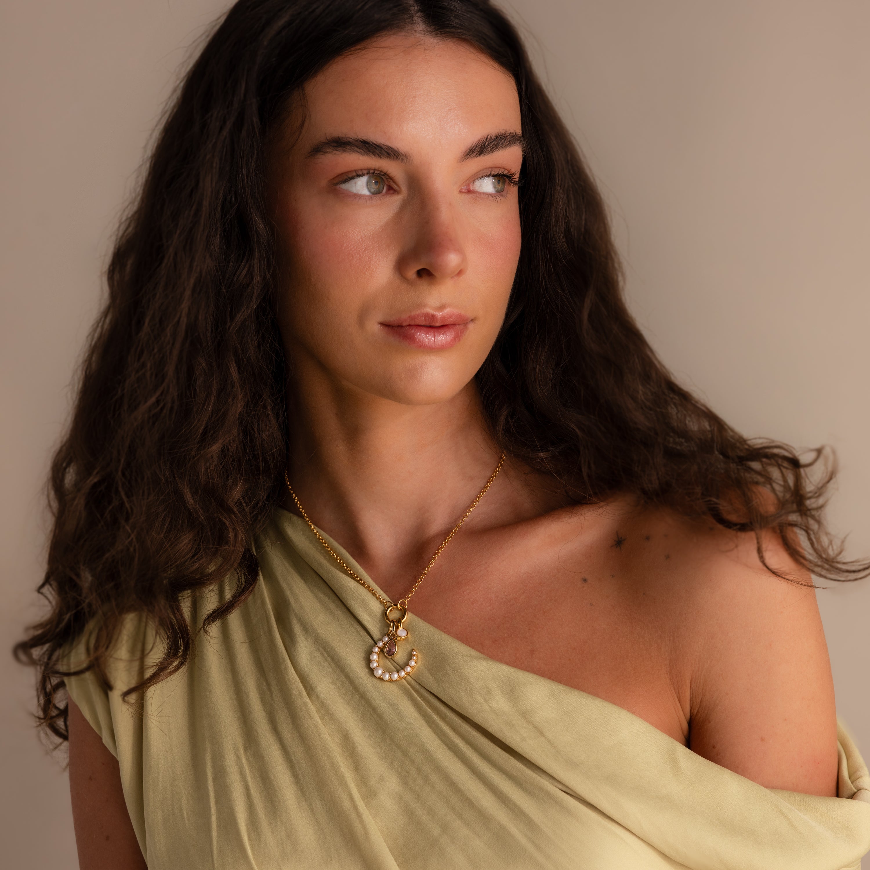 A woman with long dark hair wears a light yellow draped top and a gold necklace featuring the Pearl Moon Charm, gazing to the side—a celestial vision.