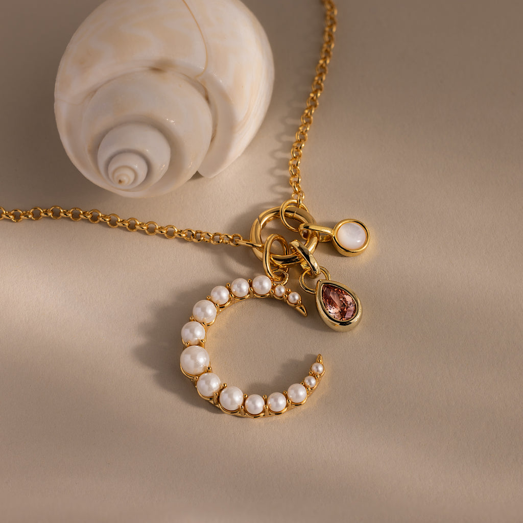 The Pearl Moon Charm necklace features celestial-inspired gemstone and pearl charms, beautifully displayed next to a white seashell on beige.