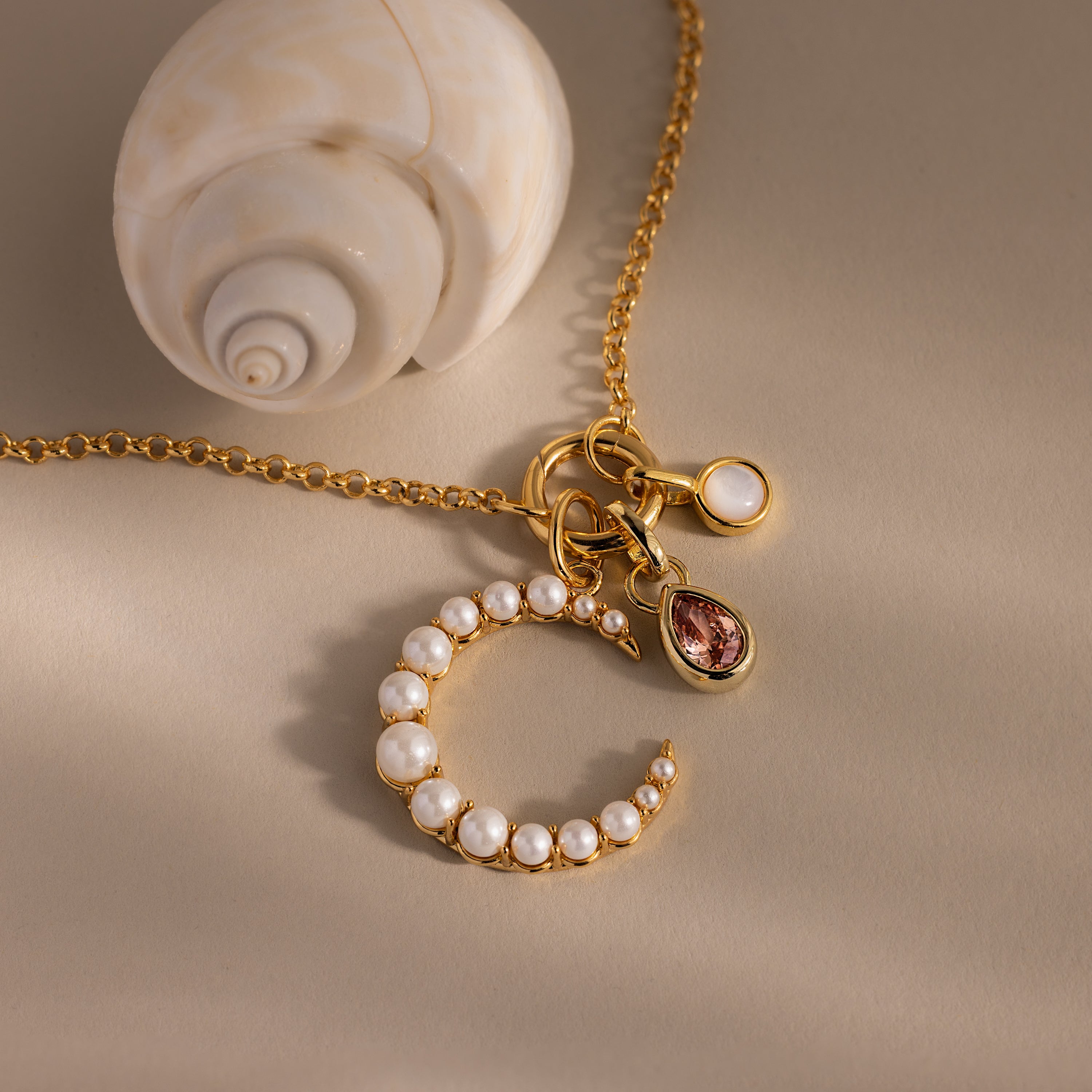 The Pearl Moon Charm necklace features celestial-inspired gemstone and pearl charms, beautifully displayed next to a white seashell on beige.