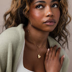 A woman with wavy hair wears the Organic Heart Locket Necklace and a cream sweater, gazing upward with a soft expression—an elegant keepsake piece.
