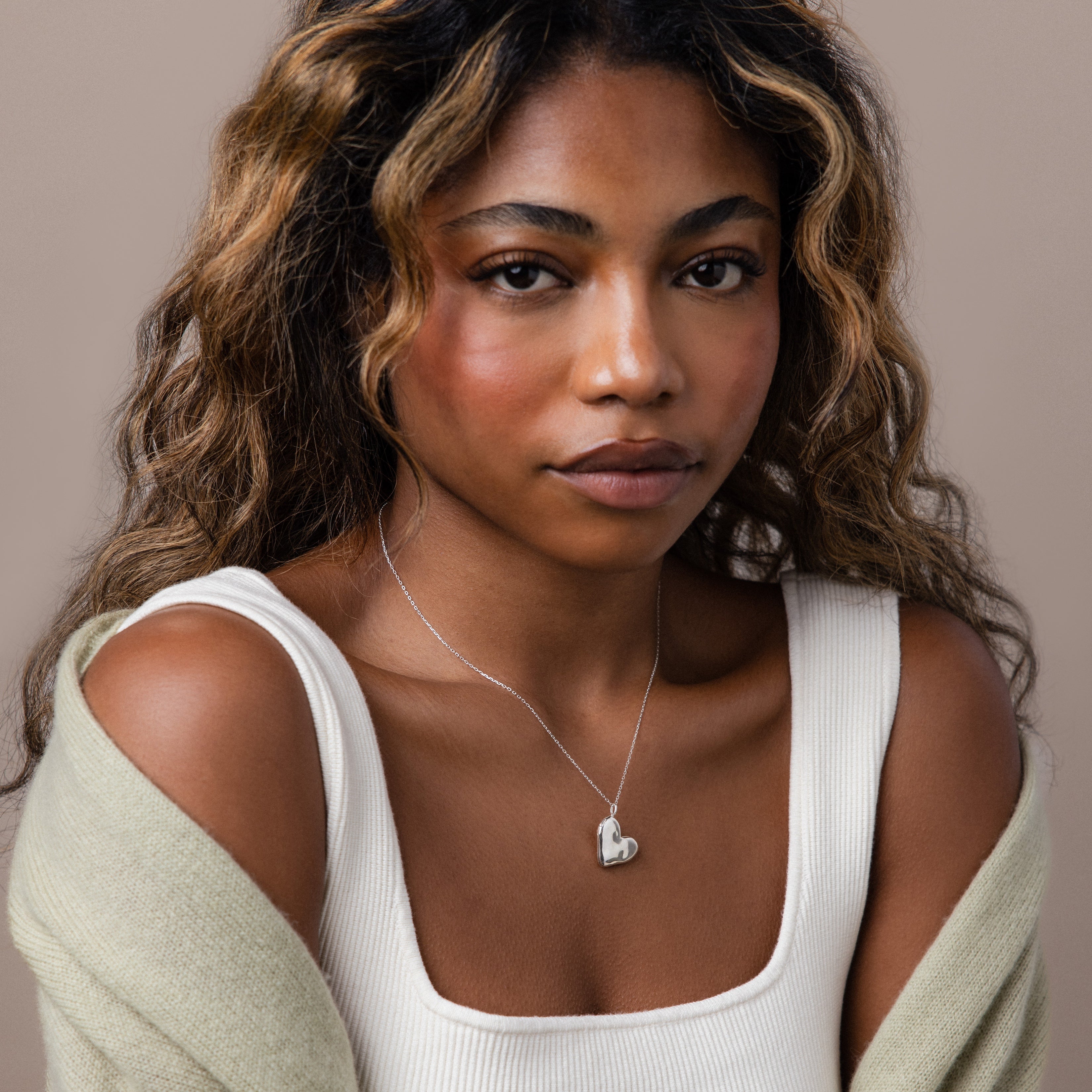 Woman with wavy hair wearing a white top, light sweater, and the Organic Heart Locket Necklace in Sterling Silver—a timeless keepsake jewelry piece.