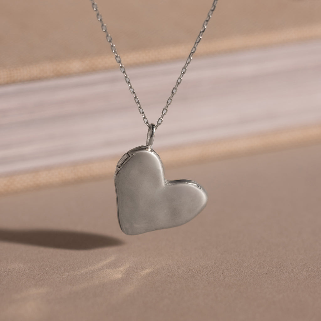 The Organic Heart Locket Necklace in Sterling Silver rests on a beige surface with a blurred book in the background, making it an ideal keepsake for preserving treasured memories.