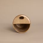 The Everyday Ceramic Mug is round and beige with a half-covered top, oval cutout handle, and subtle speckled glaze, shown on a neutral background.