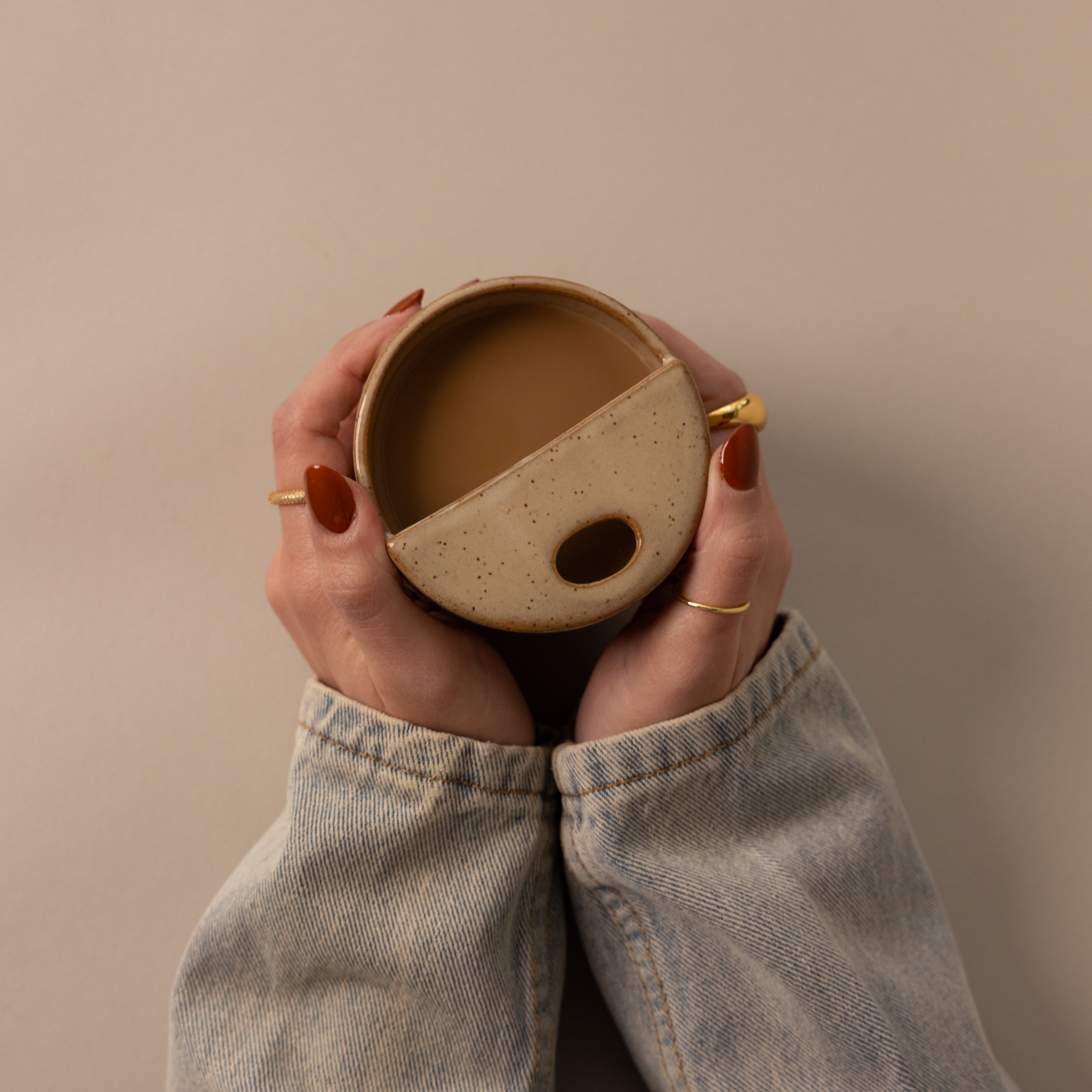Hands with painted nails hold the Everyday Ceramic Mug, featuring a speckled glaze, against a beige background while wearing a denim jacket.