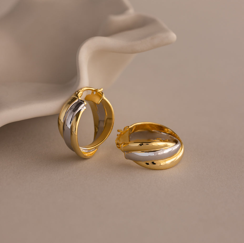 The Intertwined Mixed Metal Hoops, shown in gold and silver beside a white ceramic dish on a neutral background, bring effortless style and modern flair to any outfit.
