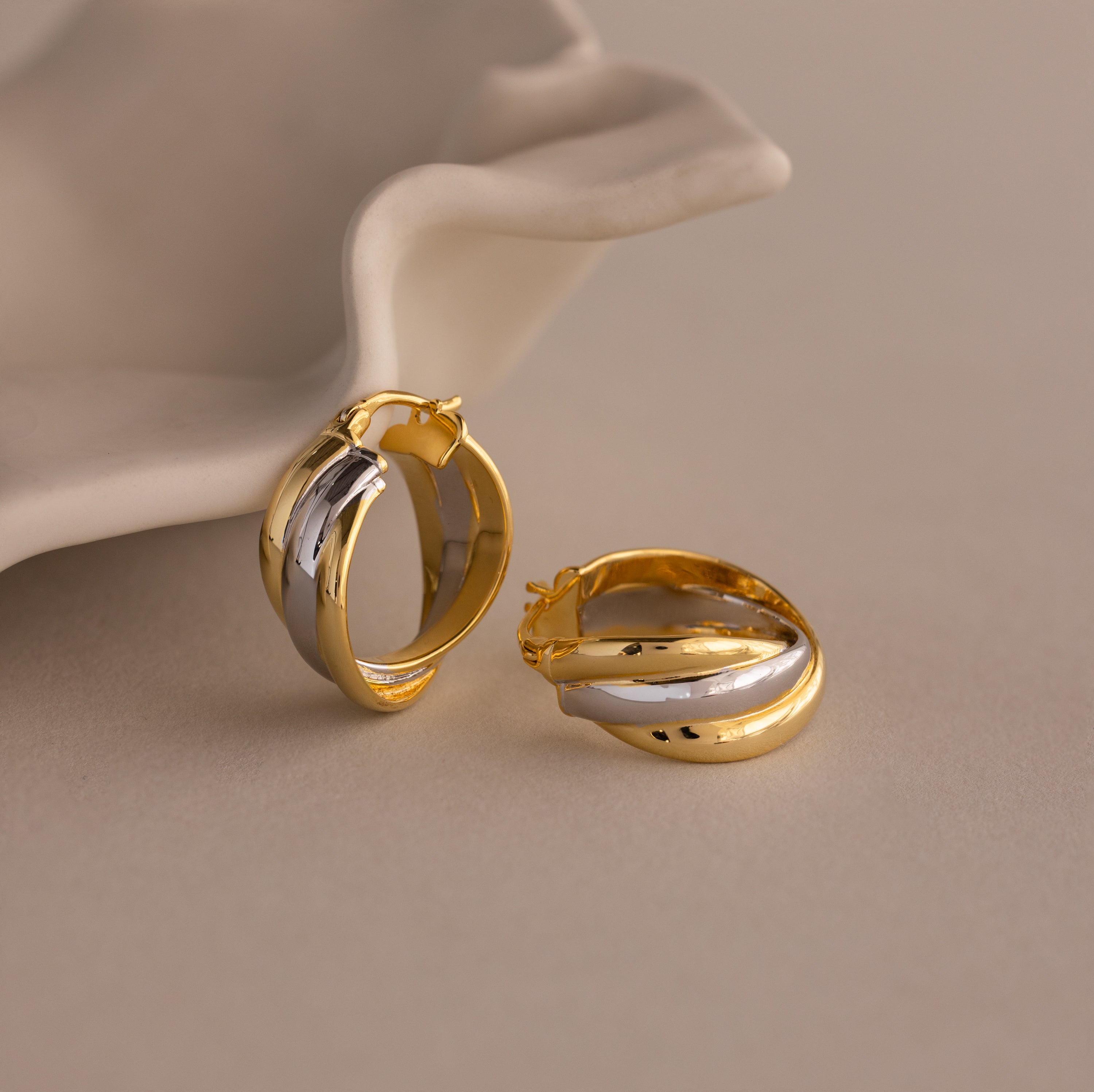 The Intertwined Mixed Metal Hoops, shown in gold and silver beside a white ceramic dish on a neutral background, bring effortless style and modern flair to any outfit.