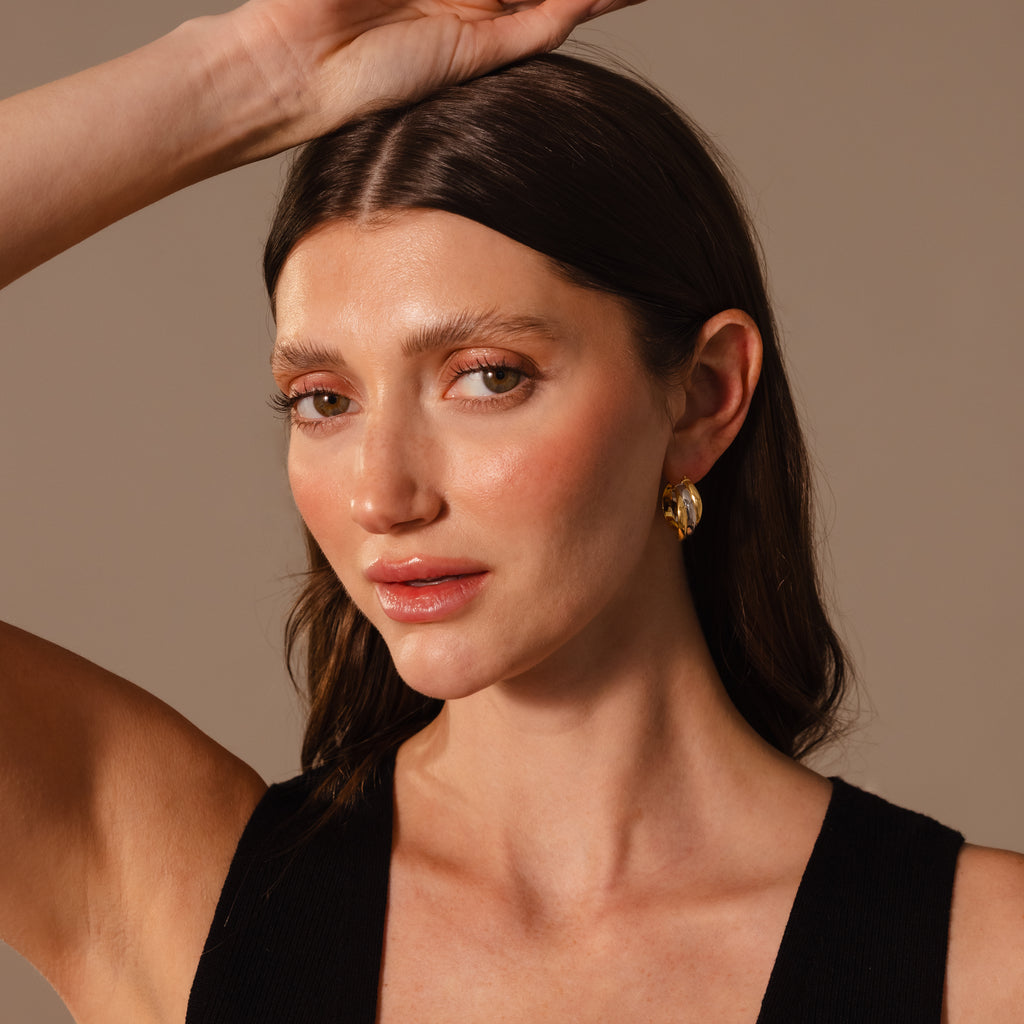 A woman with long brown hair, wearing Intertwined Mixed Metal Hoops and a black top, poses with one arm raised against a neutral background.