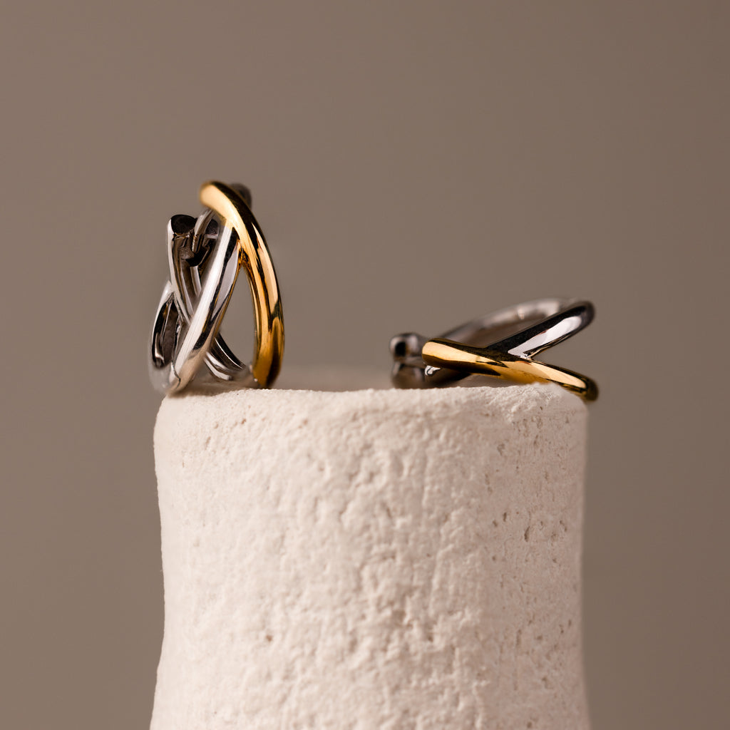 Laced Mixed Metal Huggies, featuring intertwined silver and gold in a sculptural crossover design, are showcased on a textured off-white cylindrical stand.