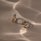 The Laced Mixed Metal Huggies, featuring gold and silver tones, are displayed on a beige surface with wavy light reflections, highlighting their sculptural crossover design.