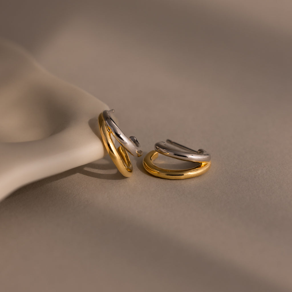 The Mixed Metal Duo Hoops—two intertwined gold and silver earrings—are displayed on a soft beige and cream surface, highlighting the contemporary elegance of these stylish hoop earrings.