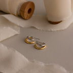 The Mixed Metal Duo Hoops, featuring gold and silver tones, are showcased on cream fabric with wooden spools in the background, highlighting a modern and contemporary style.