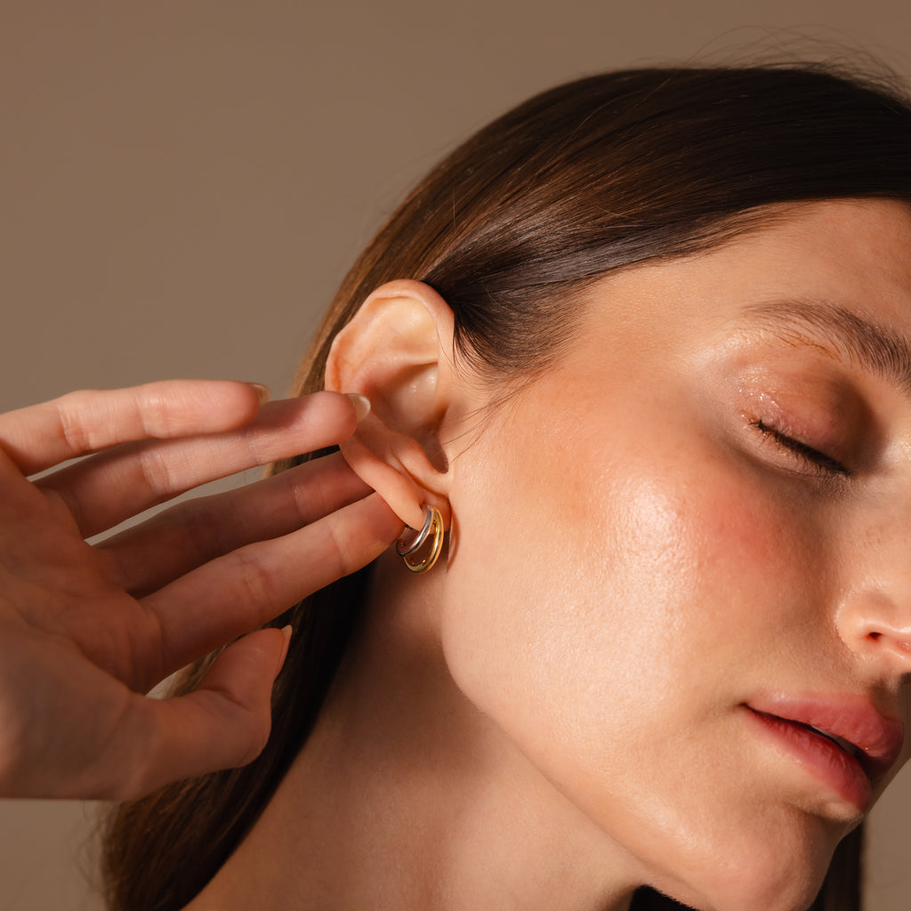 A woman with closed eyes touches a Mixed Metal Duo Hoop earring, showcasing the collection's gold and silver design against a neutral background.