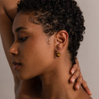 A woman with short curly hair touches her neck and looks downward in soft lighting while wearing Tiger's Eye Dome Drop Earrings.