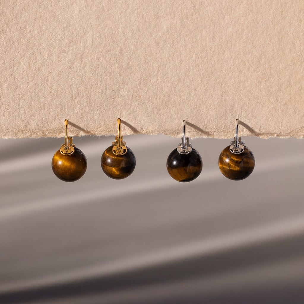 Two pairs of Tiger's Eye Dome Drop Earrings—crafted with natural stone and featuring gold or silver hooks—are elegantly displayed on a beige surface.