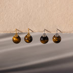 Two pairs of Tiger's Eye Dome Drop Earrings—crafted with natural stone and featuring gold or silver hooks—are elegantly displayed on a beige surface.