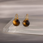The Tiger's Eye Dome Drop Earrings are elegant gold earrings featuring round Tiger’s Eye stones, beautifully presented on a clear glass surface.