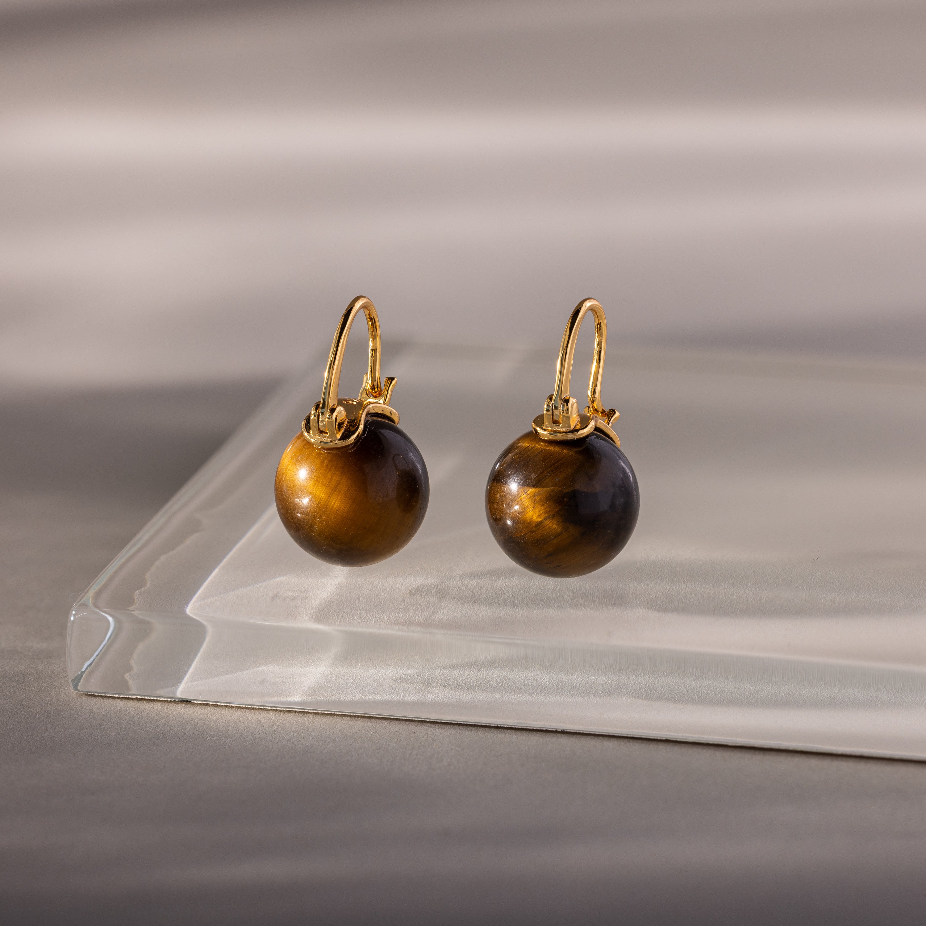 The Tiger's Eye Dome Drop Earrings are elegant gold earrings featuring round Tiger’s Eye stones, beautifully presented on a clear glass surface.