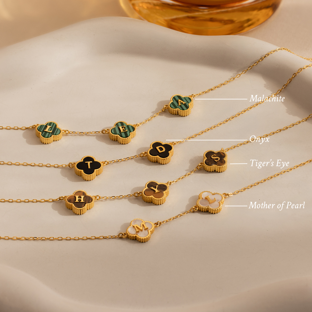 Gemstone Clover Letter Necklace in 925 sterling silver shown beside gold chain bracelets with labeled malachite, onyx, tiger’s eye, and mother of pearl charms, all displayed on a cream surface.