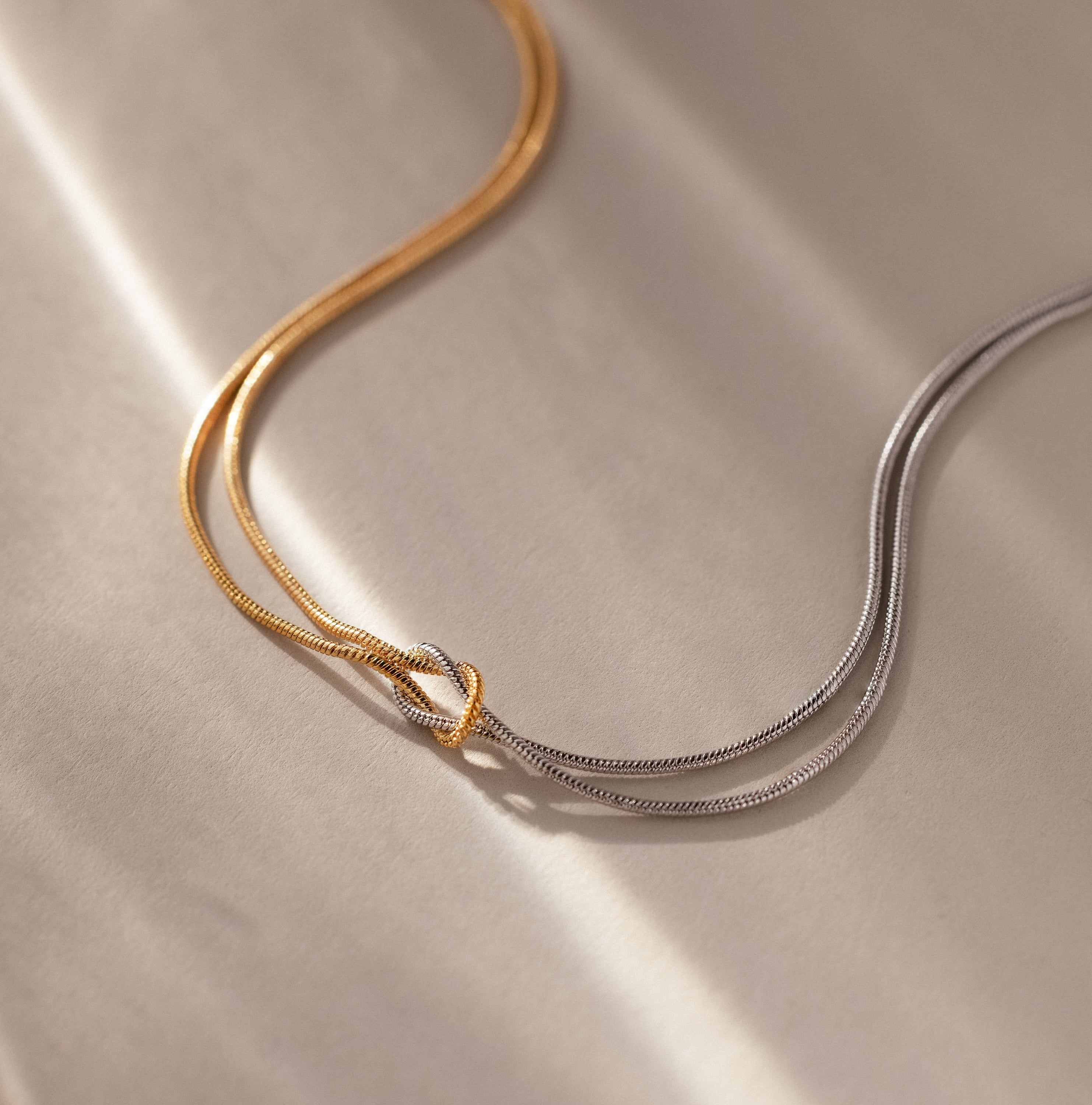 A Love Knot Chain Necklace in Mixed Metal, featuring intertwined gold and silver chains, rests gracefully on a soft, neutral background.