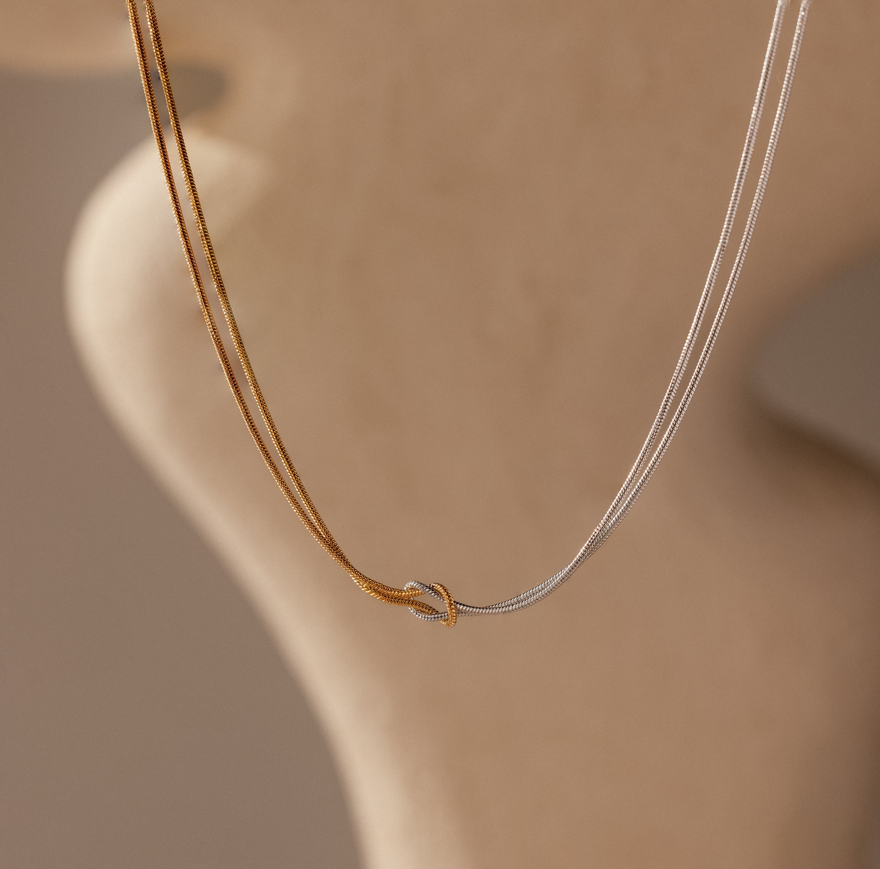 The Love Knot Chain Necklace in Mixed Metal features two intertwined gold and silver chains, displayed gracefully against a blurred beige background.