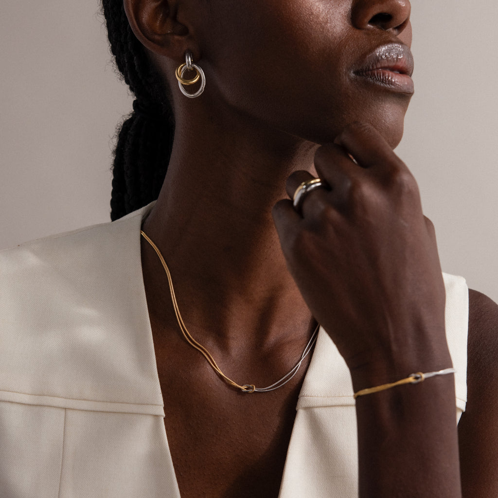 Woman wearing gold and silver jewelry, featuring the Love Knot Chain Necklace in Mixed Metal, along with matching earrings, ring, and bracelet, styled with a white sleeveless top.