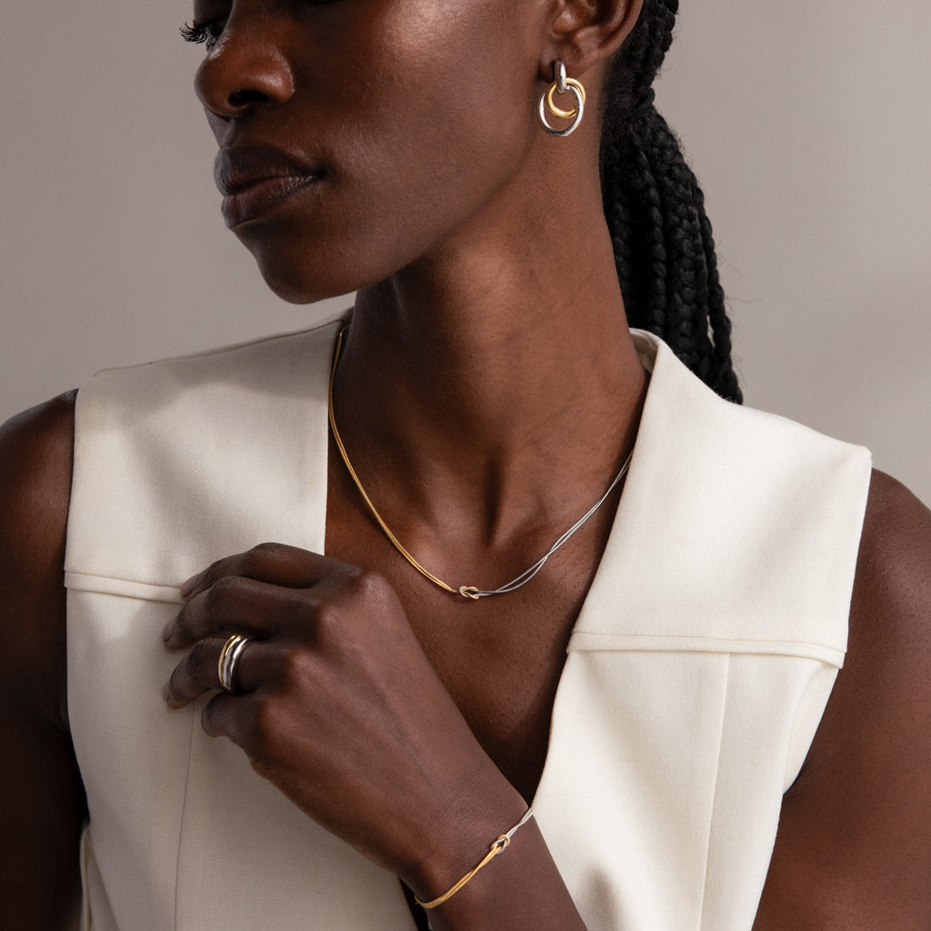 A woman with braided hair wears a Love Knot Chain Necklace in Mixed Metal and a sleeveless white top, posed against a neutral background.