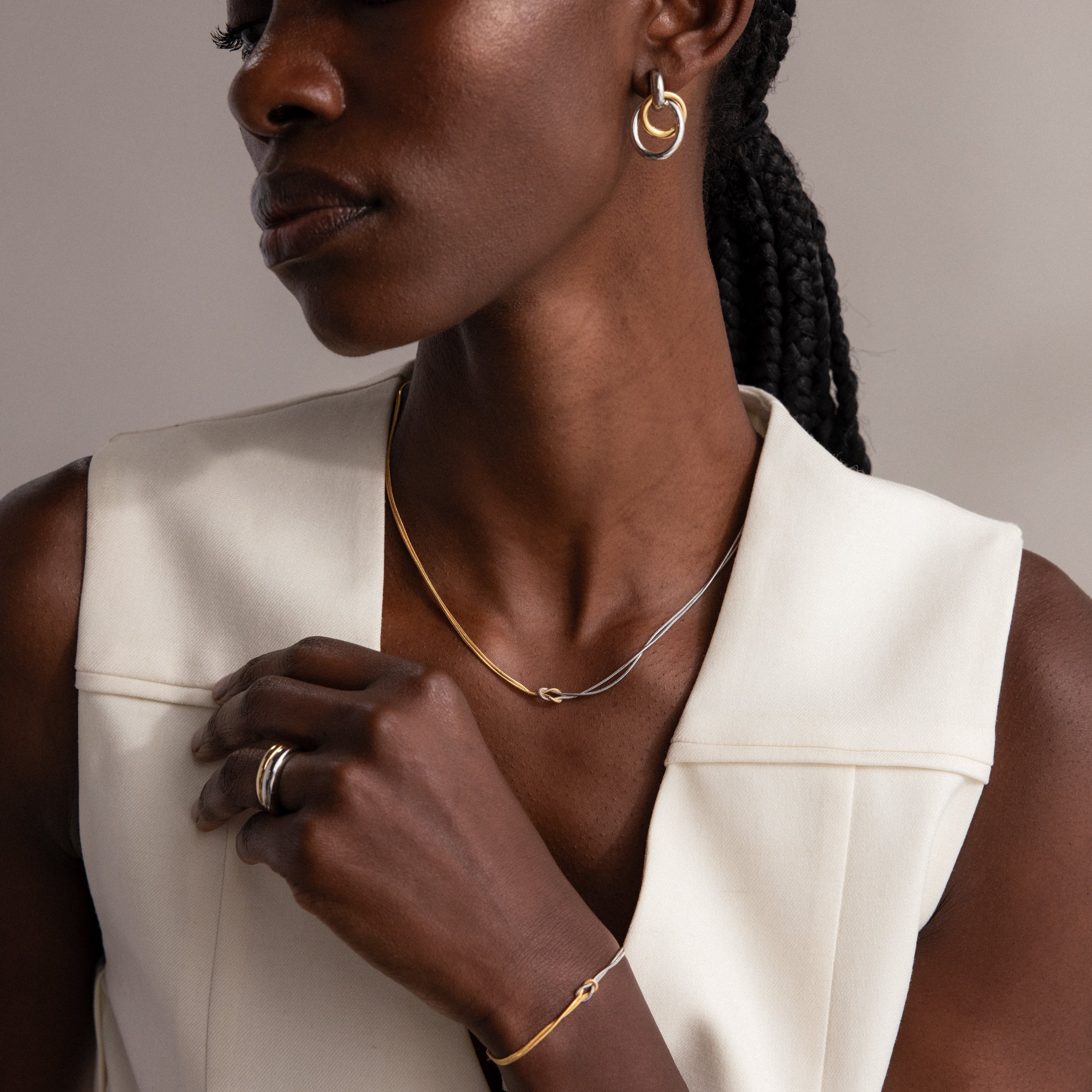 A woman with braided hair wears a Love Knot Chain Necklace in Mixed Metal and a sleeveless white top, posed against a neutral background.