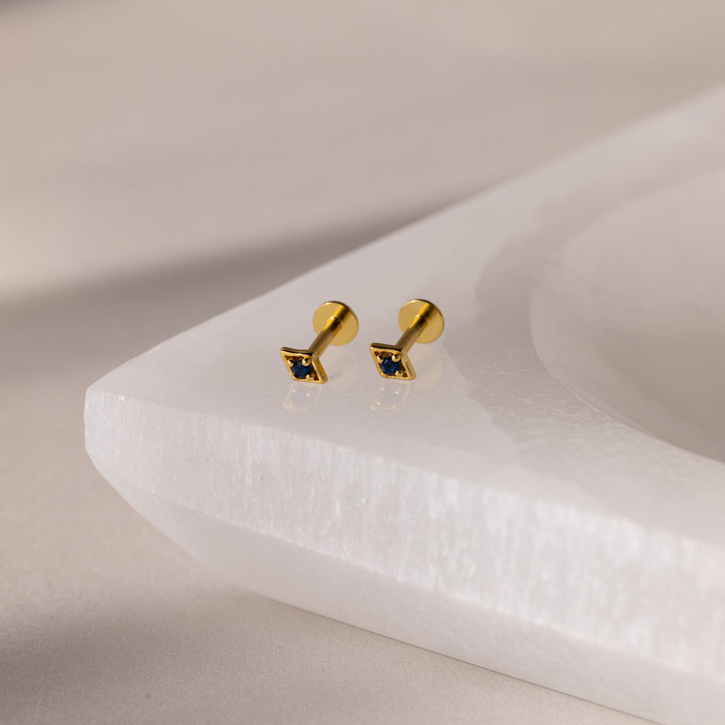 Gold geometric flat back sapphire stud earrings on a white stone surface