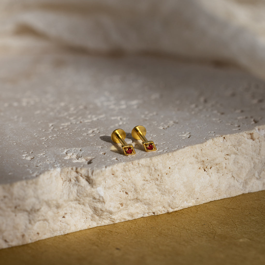 A pair of our geometric flat back garnet stud earrings with red gemstones, displayed on a textured stone surface with soft natural lighting.