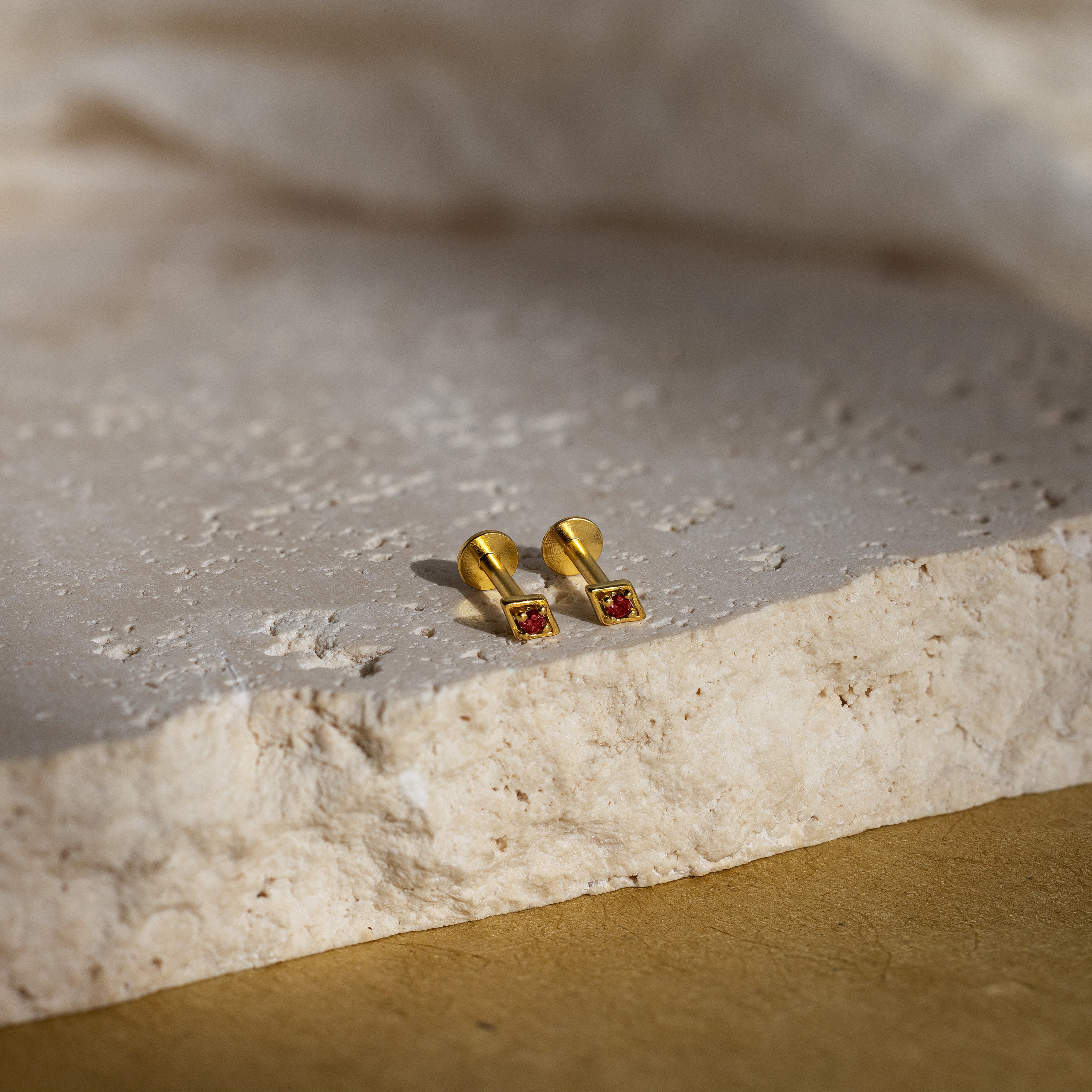 A pair of our geometric flat back garnet stud earrings with red gemstones, displayed on a textured stone surface with soft natural lighting.