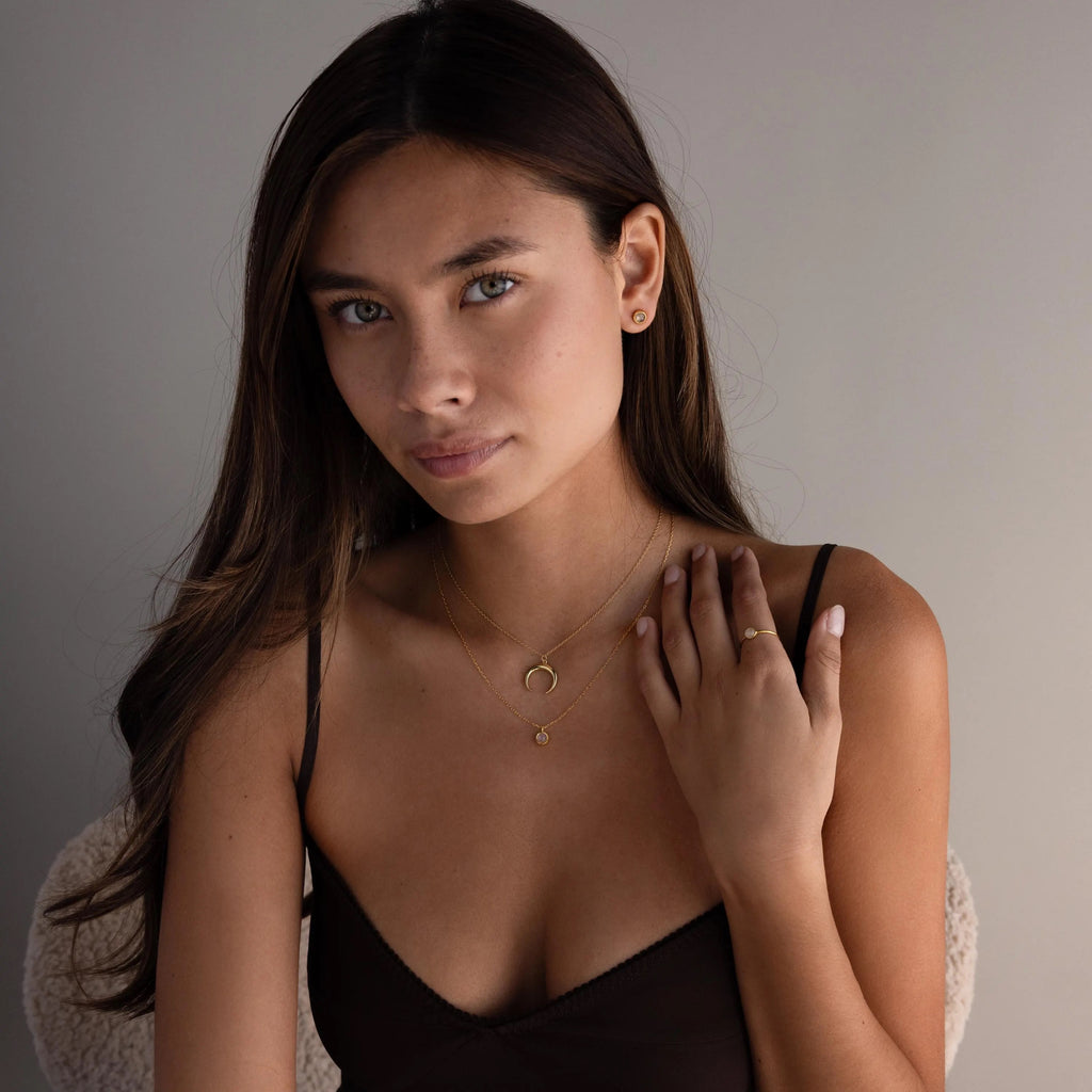 A woman with long brown hair wears a dark camisole and gold jewelry, including the Moonstone Charm Necklace, as she poses against a neutral background.