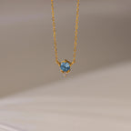 The Dainty Birthstone Drop Necklace features a gold chain with a blue gemstone pendant and a small clear stone, elegantly displayed against a neutral background.